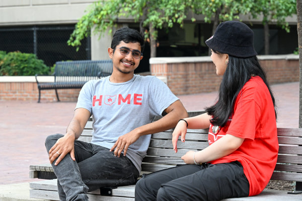 Two people sit on a wooden bench outdoors, surrounded by trees and other benches.